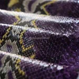 Puede incluir: Un material texturizado con un patrón geométrico repetitivo de escamas superpuestas en tonos morado intenso, amarillo brillante y gris apagado. La superficie tiene un acabado brillante con pliegues visibles que crean profundidad y sombras.