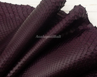 May include: A deep purple snakeskin leather sheet with a distinct, raised scale texture. The material is folded, creating soft curves and shadows across the surface. The text AcalapatiBali is printed in the centre of the dark purple hide.
