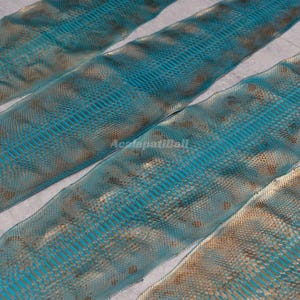 Peut inclure: Trois bandes allongées de cuir de serpent bleu sarcelle et or métallique disposées en diagonale. Le matériau présente un motif d'écailles répétitif distinct avec une teinte bleu-vert vibrante sur une base dorée texturée.