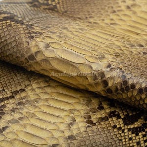 Peut inclure: Une feuille pliée de cuir de peau de serpent texturé présentant un motif naturel d'écailles superposées dans des tons jaune doré, beige et brun foncé. La surface affiche une texture d'écailles reptiliennes avec le filigrane Acalapatibali.