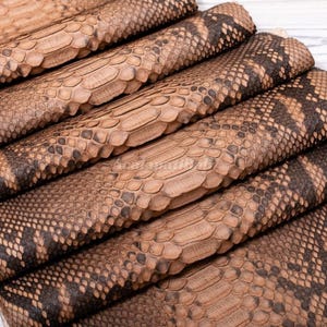 Peut inclure: Une pile de feuilles de cuir de serpent pliées, dans des tons beige et marron, présentant un motif d'écailles répétitif et distinct. La texture se compose de grandes écailles centrales allongées bordées d'écailles plus petites.