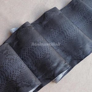 Può includere: Una serie di pannelli sovrapposti grigio antracite scuro caratterizzati da una distinta trama a pelle di serpente. Il materiale mostra un motivo a scaglie ripetuto sulla superficie, con il testo AcalopatiBali visibile al centro.
