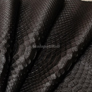 Puede incluir: Un material texturizado de color marrón oscuro con un patrón de escamas superpuestas que se repite en toda la superficie. El material está doblado, creando sombras y reflejos profundos. El texto AcalapatiBali está impreso en el centro.
