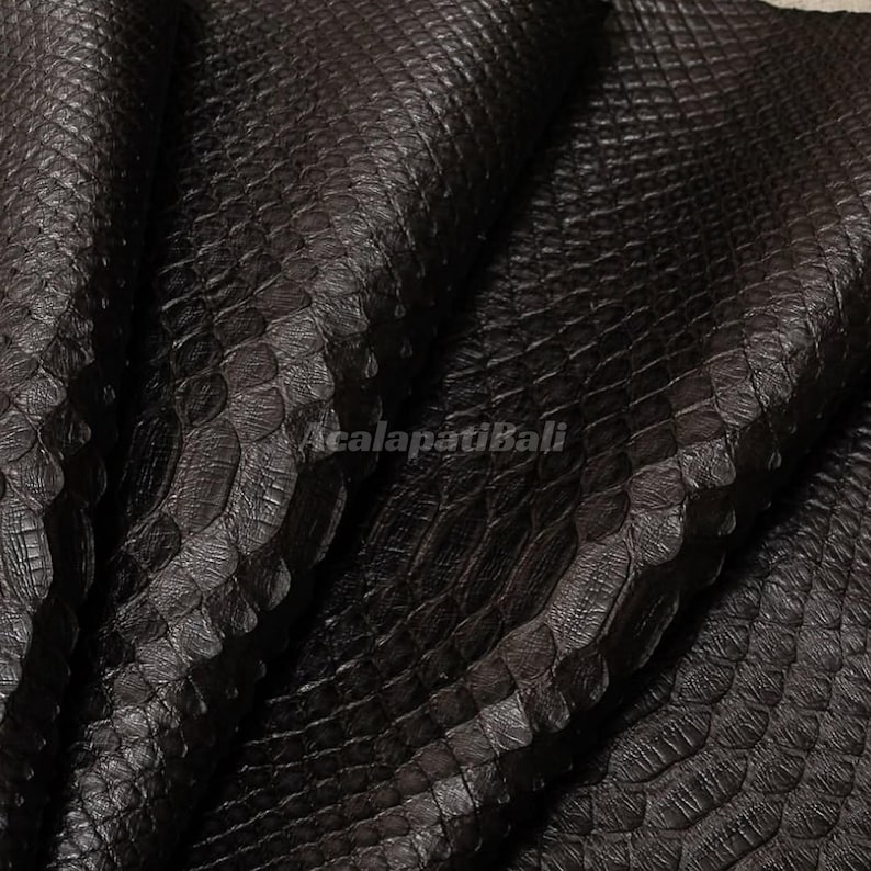 May include: A dark brown, textured material featuring a repeating, overlapping scale pattern across the entire surface. The material is folded, creating deep shadows and highlights. The text AcalapatiBali is printed in the centre of the image.