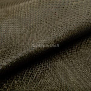 May include: A dark olive green material featuring a repeating, textured scale pattern across the entire surface. The fabric displays a folded edge, creating depth and shadows. The text AcalapatiBali is printed in a light color across the center.