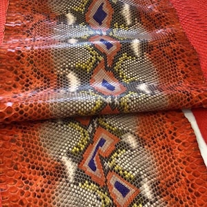 Peut inclure: Une feuille de cuir de serpent texturé présentant une couleur de base orange vif avec un motif géométrique répétitif dans des tons de jaune, gris, blanc et bleu profond. Le matériau possède une texture de surface écailleuse distincte.