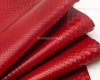 May include: A folded piece of vibrant red snakeskin leather featuring a distinct, raised scale texture throughout the surface. The material has a glossy finish and includes the text AcalapatiBali printed in the centre of the folds.