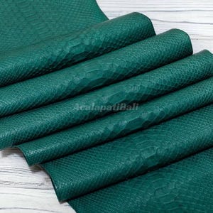 May include: A folded piece of emerald green leather featuring a textured snakeskin pattern. The surface displays a repeating scale design with a matte finish. The text AcalapatiBali is printed in a light colour across the centre of the material.