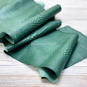May include: A piece of emerald green snakeskin leather featuring a distinct, raised scale texture across the surface. The material is folded in layers, displaying a consistent deep green colour and a repeating, natural pattern throughout the hide.