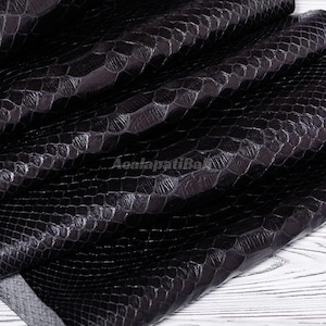 May include: A dark brown, glossy material featuring a textured snakeskin pattern with raised, overlapping scales. The surface has a reflective sheen and includes the text AcalapatiBak printed in a subtle, semi-transparent font across the centre.