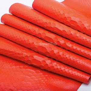 Puede incluir: Un material de cuero naranja vibrante que presenta un patrón de escamas en relieve distintivo en toda su superficie. El material está doblado en varias capas horizontales, mostrando la textura uniforme y el color brillante de la piel.