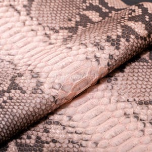 May include: A textured material featuring a repeating snakeskin pattern in shades of light pink, beige, and dark gray. The surface displays overlapping scale-like shapes with a distinct fold running diagonally across the center of the material.