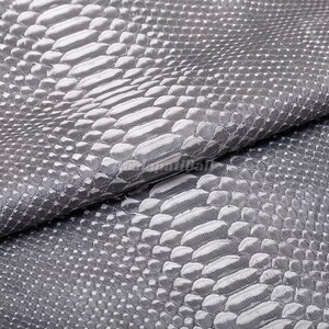 Op de afbeelding: Een vel grijs materiaal met een gestructureerd slangenleerpatroon met verhoogde, overlappende schubben. Het oppervlak toont een herhalend, organisch schubbenontwerp in verschillende tinten koel grijs, wat zorgt voor een tastbaar uiterlijk.