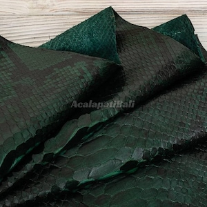 Peut inclure: Une peau de cuir de serpent vert foncé présentant un motif d'écailles distinct avec des accents noirs. Le matériau a une surface texturée et une finition brillante. Le texte AcalapatiBali est imprimé au centre du cuir plié.