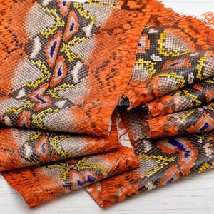 Può includere: Un pezzo di pelle di serpente piegato con un motivo vivace di squame arancioni, gialle, bianche, nere e blu. Il materiale presenta un design geometrico ripetitivo con squame testurizzate e sovrapposte su tutta la superficie.
