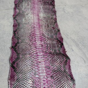 May include: A long, flat strip of snakeskin leather featuring a vibrant magenta and white gradient pattern with dark black scale outlines. The texture displays a repeating diamond-shaped scale structure running vertically along the entire length.