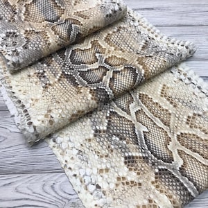 May include: A piece of textured material featuring a realistic snakeskin pattern in shades of beige, tan, brown, and white. The surface displays a repeating scale design with varying dark and light tones, creating a natural, organic appearance.