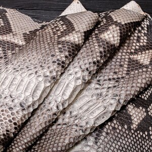 May include: A piece of textured snakeskin leather featuring a repeating pattern of overlapping scales in shades of cream, beige, gray, and dark brown. The material is folded, showcasing the intricate, natural scale structure and the varied color gradient.