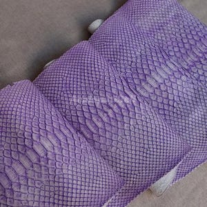 以下が含まれることがあります： ヘビ革の質感を模した、隆起した鱗模様が特徴の紫色の長方形の素材。表面は一貫したバイオレットの色合いで、鱗の間には明るく繊細な白いアンダートーンが見られます。