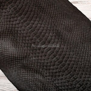 May include: A dark brown material featuring a textured, embossed pattern that mimics the appearance of reptile scales. The surface displays a repeating, organic scale design with a matte finish and the text Acalapatibali visible in the centre.