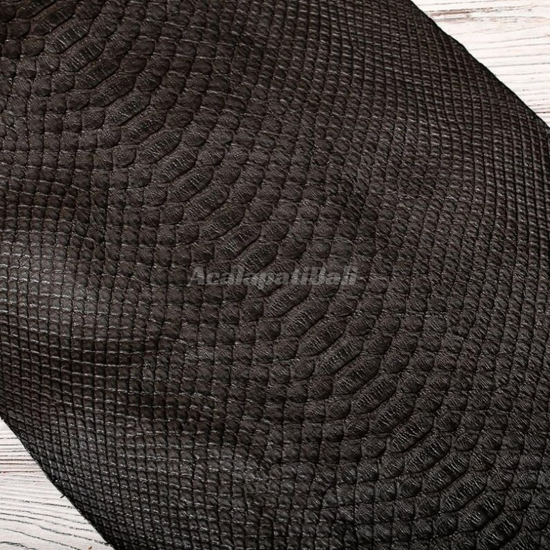 May include: A dark brown material featuring a textured, embossed pattern that mimics the appearance of reptile scales. The surface displays a repeating, organic scale design with a matte finish and the text Acalapatibali visible in the centre.