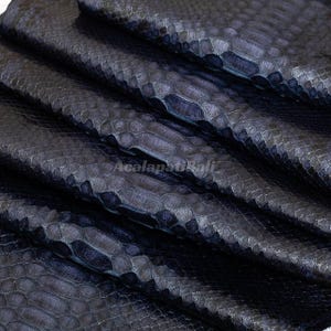 Puede incluir: Una piel de serpiente de color azul marino oscuro con una textura de escamas realzada y distintiva. La superficie presenta un patrón repetitivo de escamas superpuestas e incluye el texto de marca de agua AcalapatiBali en el centro.