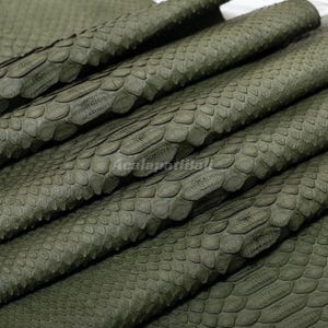Peut inclure: Une feuille de cuir vert olive présentant un motif gaufré texturé imitant l'apparence d'écailles de serpent. Le matériau est plié en plusieurs couches, mettant en valeur le motif répétitif des écailles et la finition mate de la surface.