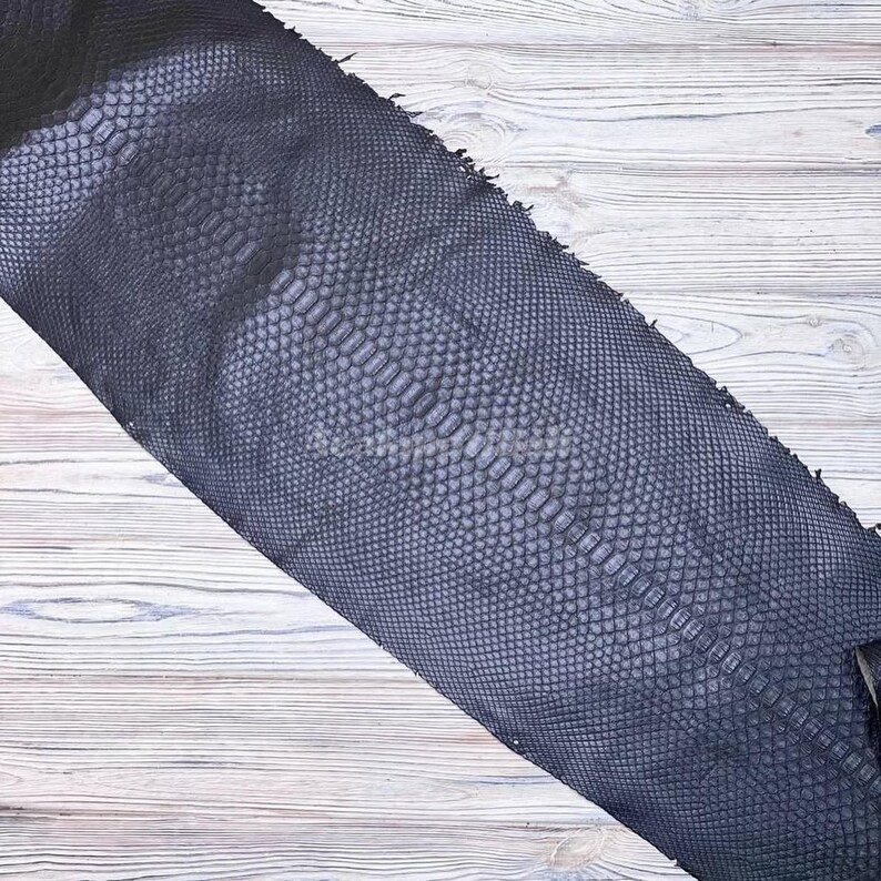 May include: A dark navy blue snakeskin leather hide featuring a distinct, repeating scale pattern across the entire surface. The material displays a textured, organic appearance with a central row of larger, elongated scales flanked by smaller scales.