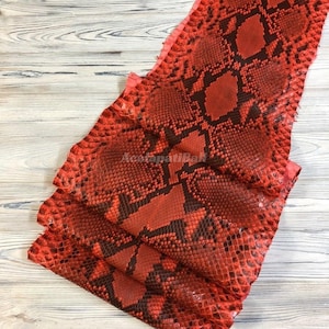 Peut inclure: Une pièce de cuir de serpent rouge et noir présentant un motif d'écailles en forme de losange. Le matériau possède une surface texturée avec une couleur de base rouge vif et des marques noires foncées créant un motif géométrique répété.