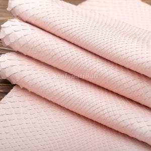 Könnte beinhalten: Ein gefaltetes Stück hellrosa Schlangenleder mit einem ausgeprägten, sich wiederholenden rautenförmigen Schuppenmuster auf der gesamten Oberfläche. Das Material hat ein mattes Finish und liegt in mehreren weichen, überlappenden Falten.