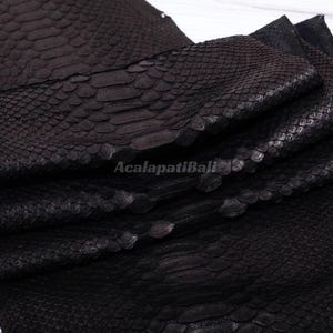 May include: A dark black snakeskin leather hide featuring a distinct, raised scale pattern across the surface. The material is folded in layers, showcasing the texture and depth of the scales. The text AcalapatiBali is visible in the center.