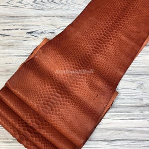 Könnte beinhalten: Ein Stück rostfarbenes Leder mit einer ausgeprägten, geprägten Schlangenhautstruktur. Das Material ist in sich gefaltet und zeigt das sich wiederholende Schuppenmuster auf der Oberfläche. Das Leder hat ein mattes Finish und einen tiefen, warmen Braunton.