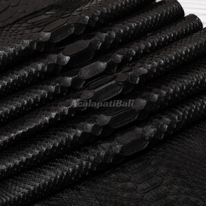 May include: A folded sheet of black snakeskin leather featuring a distinct, raised scale pattern across the surface. The material has a glossy finish and displays the text AcalapatiBali printed in a light gray font across the center.