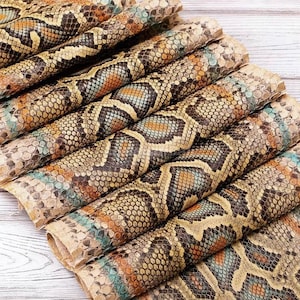Puede incluir: Una pila de hojas plegadas de material texturizado con un patrón repetitivo de piel de serpiente en tonos beige, marrón, negro y azul verdoso apagado. La superficie muestra una textura detallada de escamas superpuestas en todo el conjunto.