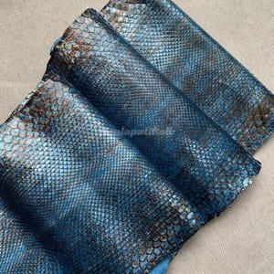 Può includere: Un pezzo di pelle testurizzata con un motivo a pelle di serpente blu metallizzato e marrone. Il materiale presenta un design a scaglie ripetute con una finitura scintillante, mostrando uno strato piegato che rivela l'intricata trama a scaglie.