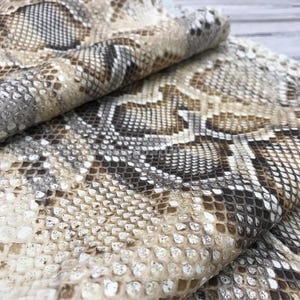 May include: A folded piece of material featuring a realistic snakeskin pattern in shades of tan, brown, white, and dark grey. The surface displays a repeating, textured scale design that mimics the appearance of natural reptile skin throughout.