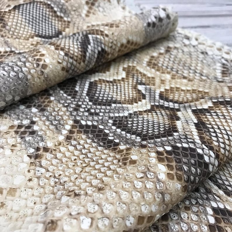 May include: A folded piece of material featuring a realistic snakeskin pattern in shades of tan, brown, white, and dark grey. The surface displays a repeating, textured scale design that mimics the appearance of natural reptile skin throughout.