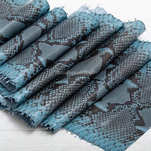 Può includere: Un pezzo di pelle testurizzata con un motivo ripetitivo a squame di serpente in tonalità turchese brillante e grigio antracite scuro. Il materiale è piegato in più strati, mostrando la texture dettagliata e sovrapposta delle squame.