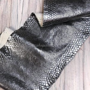 May include: A piece of metallic silver leather featuring a textured snakeskin pattern. The material has a reflective, shiny finish with a repeating scale design across the surface. The underside of the leather is visible as a light beige colour.