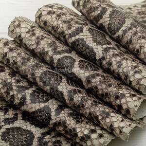 Peut inclure: Une pile de feuilles de cuir de serpent véritable pliées présentant un motif d'écailles naturel dans des tons crème, beige et brun foncé. Le matériau présente une surface texturée avec des écailles superposées disposées en motif organique.