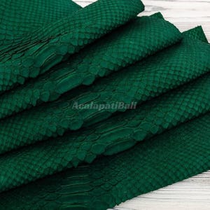 May include: A deep emerald green snakeskin leather material featuring a distinct, raised scale pattern across the surface. The material is folded in layers, displaying the texture and the text AcalapatiBali printed in the centre of the image.