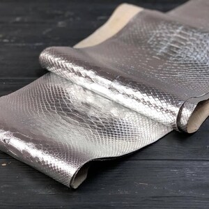 May include: A folded piece of metallic silver leather featuring a distinct embossed snakeskin texture. The material has a reflective, high-shine finish with a visible light-coloured underside along the edges, showcasing the flexible nature of the hide.