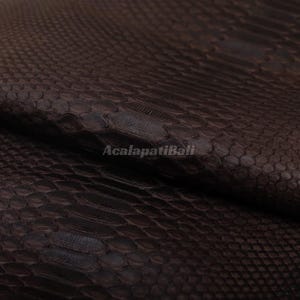 Peut inclure: Un matériau brun foncé avec un motif texturé de écailles répétitives ressemblant à de la peau de serpent. La surface présente un design à écailles hexagonales gaufrées cohérent, avec le texte AcalapatiBali imprimé en clair au centre.