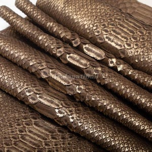 Puede incluir: Una pila de láminas superpuestas de piel de serpiente de color bronce metálico. El material presenta un patrón de escamas repetitivo y distintivo con un acabado reflectante y brillante que resalta la textura y profundidad de cada escama en la superficie.