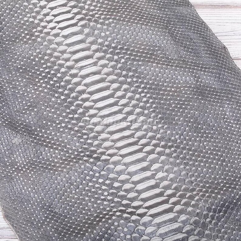 Op de afbeelding: Een vel grijs materiaal met een gestructureerd slangenleerpatroon. Het oppervlak toont een centrale rij grotere, langwerpige, rechthoekige schubben, geflankeerd door kleinere, korrelige, ruitvormige schubben over de gehele breedte.