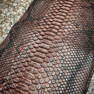 May include: A textured material featuring a repeating pattern of overlapping scales in shades of deep copper, bronze, and dark charcoal black. The surface displays a distinct, raised scale-like structure with a metallic sheen and dark edges.