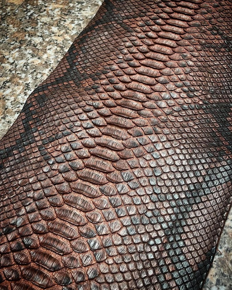 May include: A textured material featuring a repeating pattern of overlapping scales in shades of deep copper, bronze, and dark charcoal black. The surface displays a distinct, raised scale-like structure with a metallic sheen and dark edges.