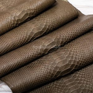 Può includere: Un pezzo piegato di pelle di serpente color tortora caratterizzato da un motivo a squame distinto e ripetitivo su tutta la superficie. Il materiale ha una finitura liscia e semilucida con una trama visibile e il testo AcalapatiBali stampato al centro.