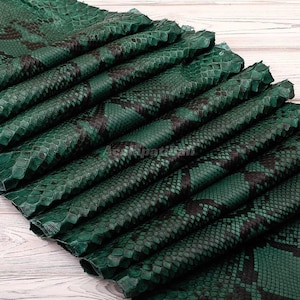 May include: A stack of folded emerald green snakeskin leather sheets featuring a dark black diamond-shaped scale pattern. The textured material is layered in overlapping sections, displaying the natural reptilian scale structure and deep green tones.