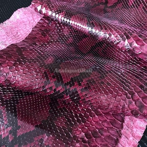 Peut inclure: Une feuille de cuir de serpent magenta et noir présentant un motif d'écailles répétitif distinct avec une finition brillante et réfléchissante. La surface affiche un dégradé de tons rose foncé et sombres, avec le texte AcalapatiBali visible.